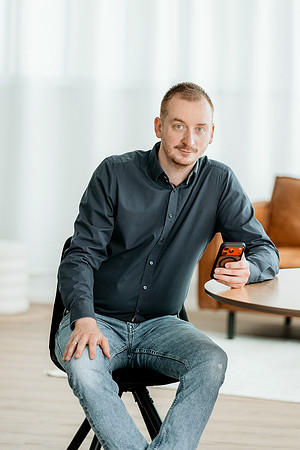 Modernes Business-Portrait für Personal Brand Fotografie im Allgäu: Ein IT-Berater sitzt lächelnd auf einem Stuhl in einem hellen Büro-Studio und hält ein Smartphone in der Hand.