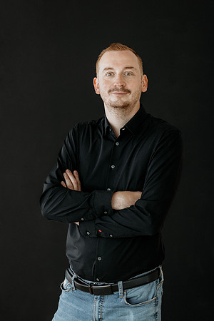 Klassisches Business-Portrait für Unternehmensfotografie in Lindau: Ein Mann in schwarzem Hemd blickt mit verschränkten Armen selbstbewusst vor einem tiefschwarzen Hintergrund in die Kamera.