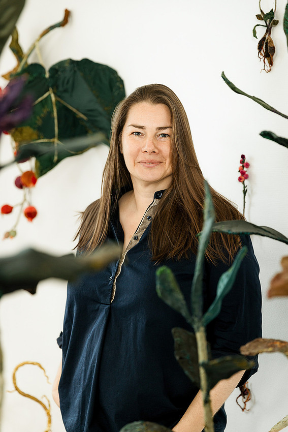 Charakterportrait von Anne Carnein: Die Künstlerin blickt entspannt durch die grünen Blätter ihrer Skulpturen im Atelier.