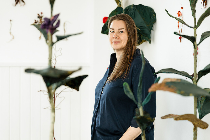 Künstlerportrait im Allgäu & Bodensee: Bildhauerin Anne Carnein steht lächelnd in ihrem Atelier neben großen Pflanzen.