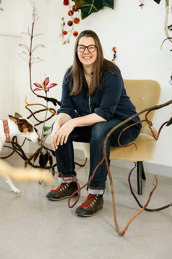 Atelierreportage im Allgäu: Künstlerin Anne Carnein sitzt lächelnd auf einem Stuhl inmitten ihrer textilen Natur-Kunstwerke.