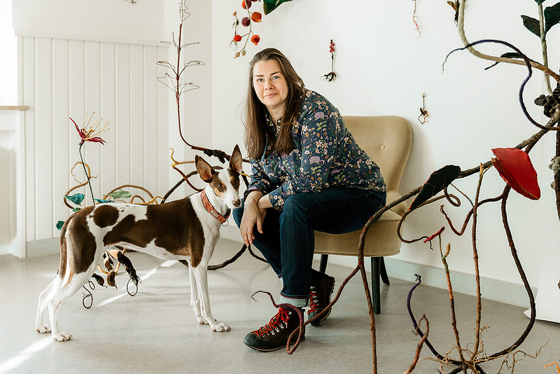 Künstlerportrait mit Hund: Anne Carnein sitzt lachend auf einem Stuhl in ihrem Atelier, neben ihr steht ihr Hund vor einer Skulptur.