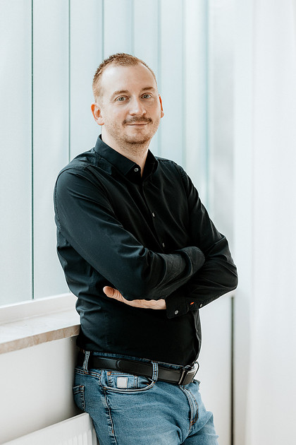 Businessfotografie Schussental Mann in schwarzem Hemd mit verschränkten Armen lehnt an heller Wand.